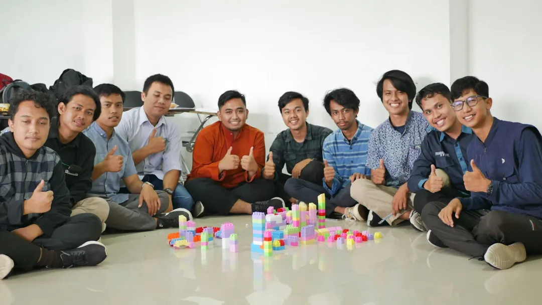 A team sits on the floor around a toy-brick build, giving a thumbs-up