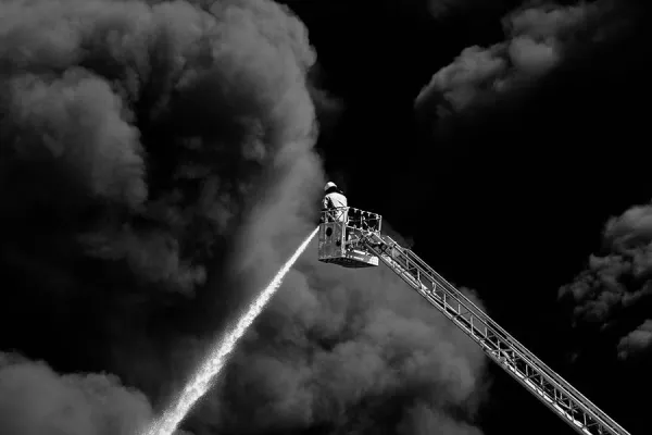 Firefighter standing on extending ladder firing a water canon into a black cloud of smoke