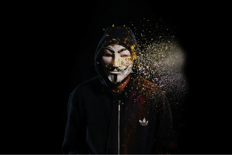 Figure wearing a Guy Fawkes mask standing in front of a black background, a burst of debris is coming from the figure's left