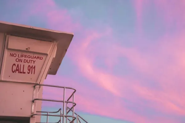 Sunset photo of a lifeguard stand with pink clouds in the background. A sign on the side says No Lifeguard on Duty, call 911.